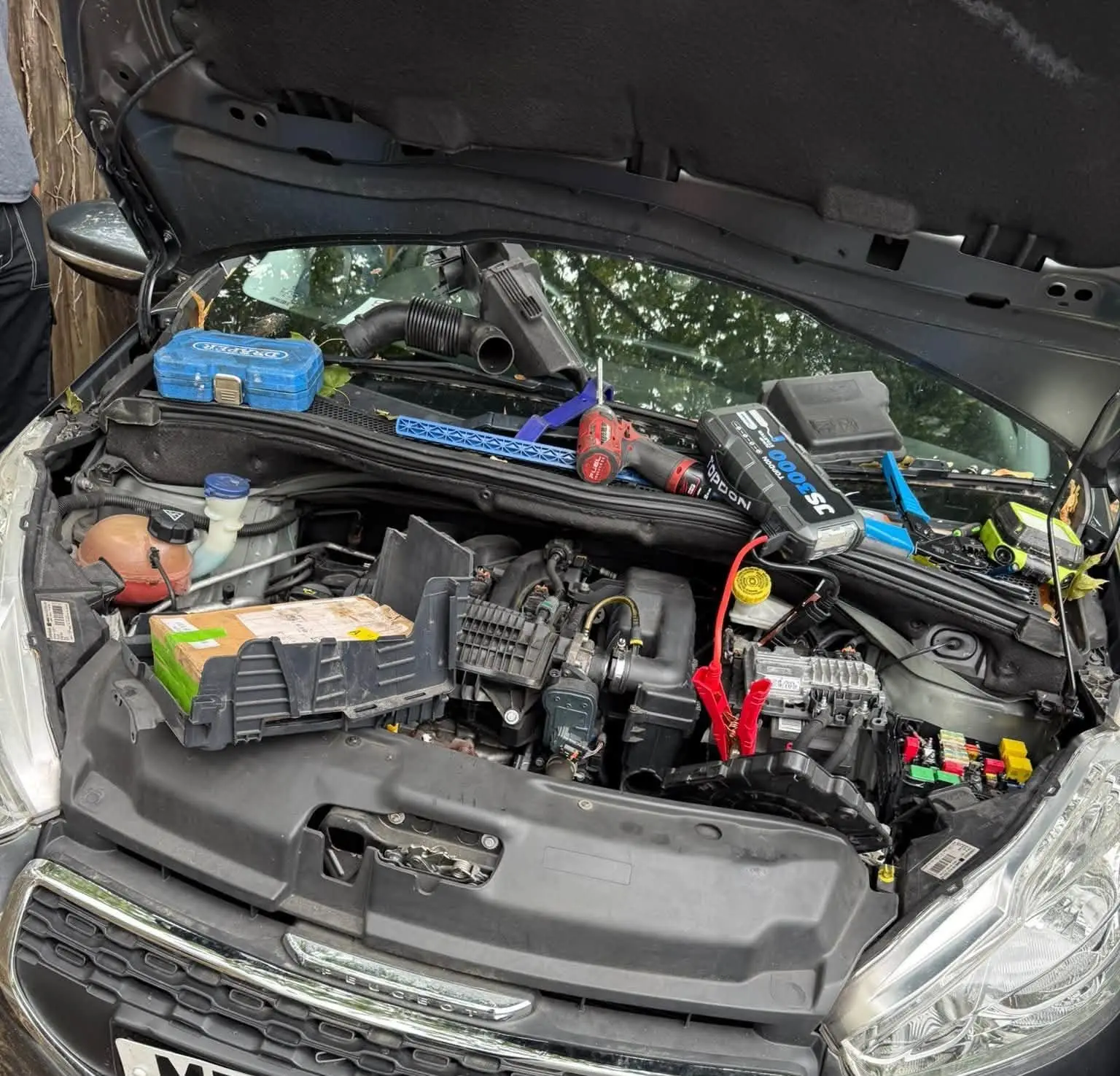 Jump pack connected under the bonnet of a hatchback with tools laid out across the engine bay