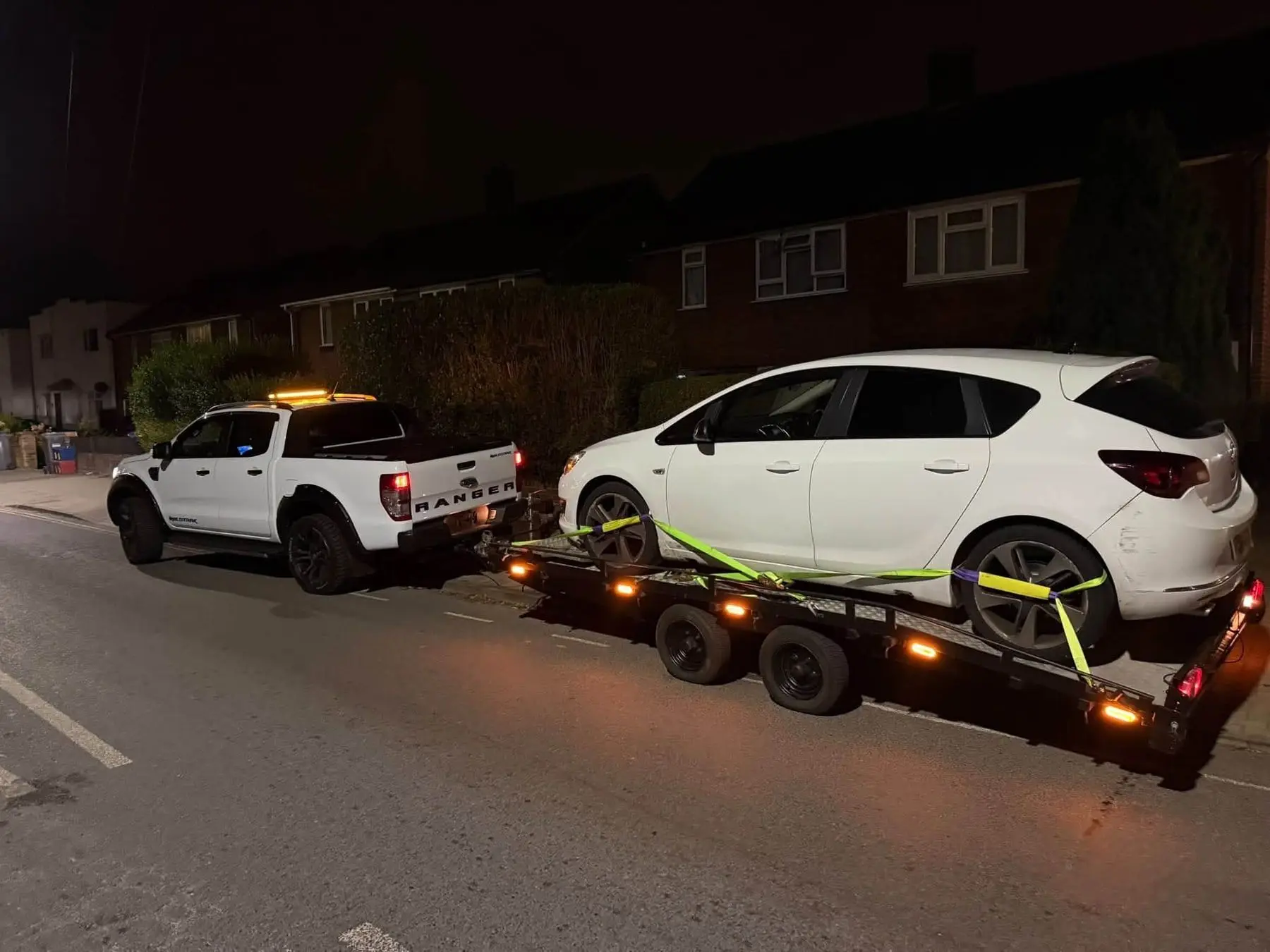The LTR Ford Ranger with its amber beacon lit, towing a hatchback strapped to the transporter trailer at night