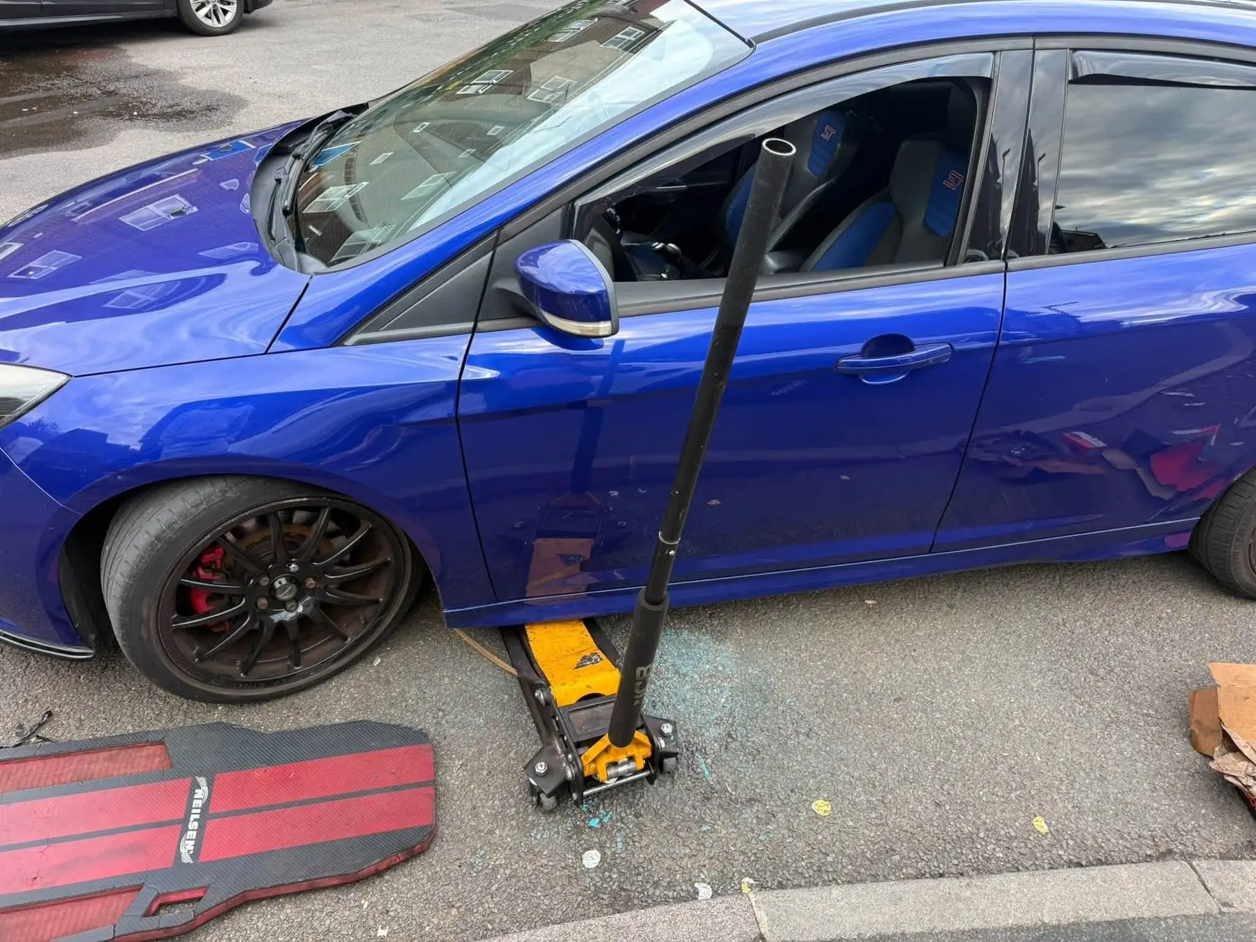 Blue hatchback raised on a trolley jack with the front wheel off for brake work